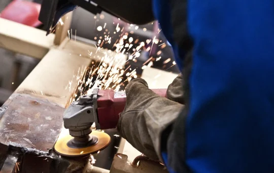 Gloved hands run an angle grinder across metal, sparks streaming off the wheel