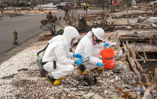 Two workers in disposable suits and respirators crouch to sample burned debris