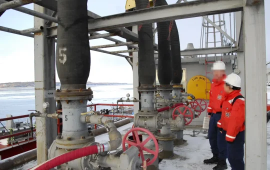 Two inspectors in hard hats stand among the valves and hoses of a bulk liquid transfer manifold