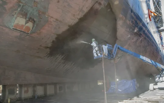 A blaster in a hood and protective suit on a boom lift abrasive-blasts a ship hull in a cloud of dust