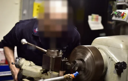 A machinist in safety glasses leans in to watch a metal part turning on a lathe