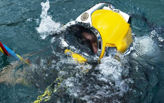 A diver in a yellow surface-supplied helmet breaks the surface of the water