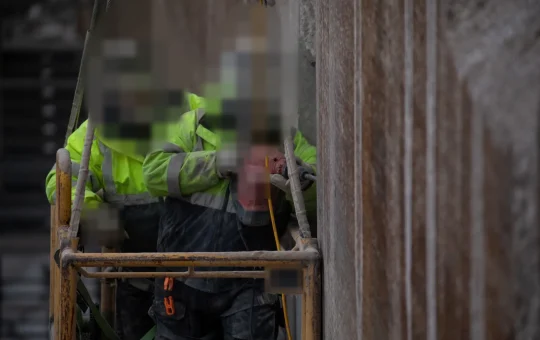Two workers ride a personnel platform hung from a crane beside a concrete lock wall