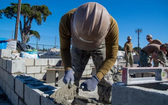 A mason in a hard hat spreads mortar on a concrete block wall