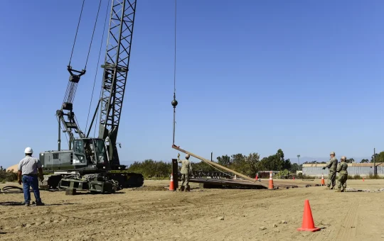 An operator in the cab of a lattice boom crawler crane lifts sheet pile while a crew holds tag lines