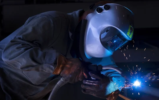 A welder in a welding helmet, leather gloves and work jacket runs a bead as sparks fly