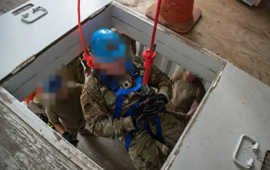 An entrant in a full-body harness on two retrieval lines climbs through a hatch while team members wait in the space below