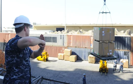 A signal person, seen from behind, gives a hand signal as a crane lifts a crate on a pier
