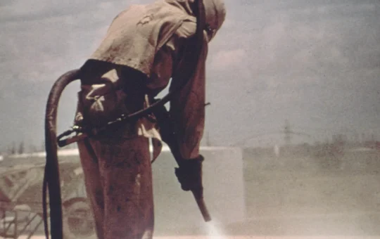 A blasting operator in a hood and full protective suit sprays abrasive at a surface