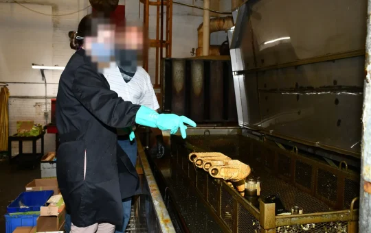 Two engineers in safety glasses and gloves look over a basket of aircraft parts in a large heated chemical stripping tank
