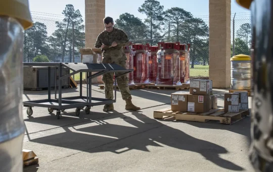 Labeled shipping boxes on a pallet beside marked drums at a receiving dock