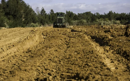 A road grader shapes a dirt road through scrub