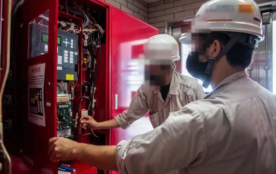 Two technicians in hard hats inspect the open control panel of a building fire alarm system