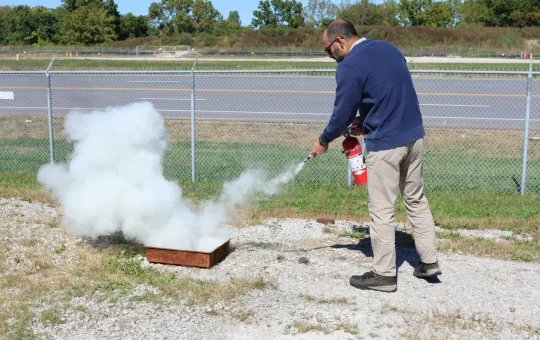 A trainee sprays a fire extinguisher at the base of a small pan fire