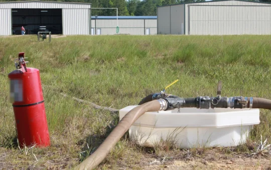 A portable fire extinguisher stands beside a fuel hose valve set over a spill containment tub at a fuel point