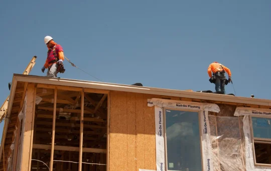 Two workers on the roof of a framed house, each tied off to a lifeline