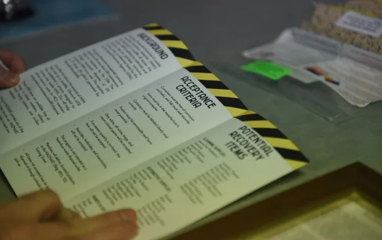 A hand holds a printed checklist in a chemical storeroom with labeled containers behind