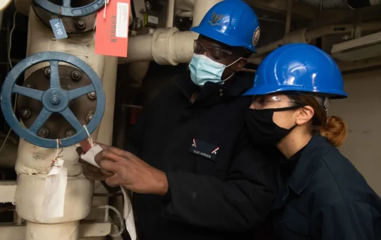 Two workers in hard hats and masks attach a tag to a valve handwheel