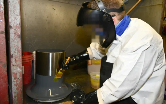 A worker in a face shield, chemical-resistant gloves, apron and coat brushes chemical paint stripper onto a metal part