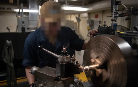 A machinist in safety glasses works at a lathe beside the spinning chuck