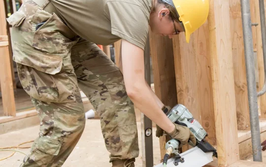 A worker in a hard hat drives a pneumatic nailer into the base of a stud wall