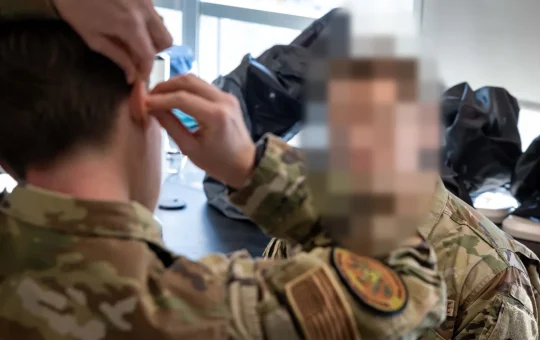 A trainee rolls a foam earplug into his ear while an instructor watches