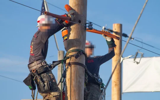 Two linemen in hard hats and harnesses work at the top of a pole