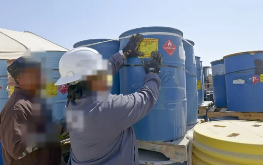 Workers fix a yellow HAZARDOUS WASTE label and a flammable liquid hazard diamond to blue drums in a waste storage yard