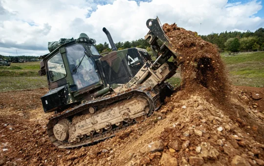 A bulldozer with an enclosed protective cab climbs a steep dirt berm