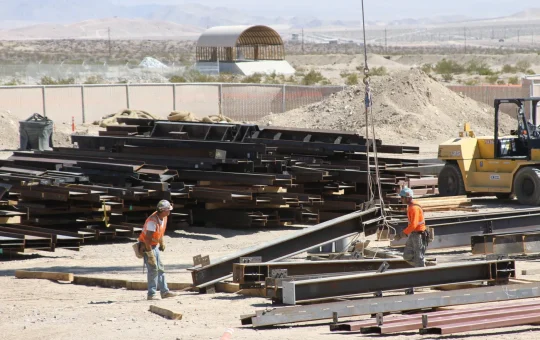 Two ironworkers among stacked steel beams as a crane line comes down to the pick