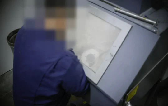 A worker sandblasts metal parts inside a closed blast cabinet, working through the glove ports and window