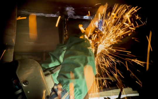 A shipyard worker in a welding hood grinds under a ship's hull, sparks flying