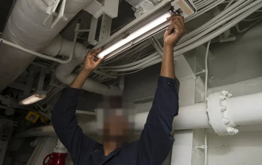A technician in safety goggles changes a tube lamp in an overhead light fixture