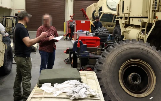 A shop worker explains something to a safety specialist holding a clipboard during an inspection of a vehicle maintenance bay
